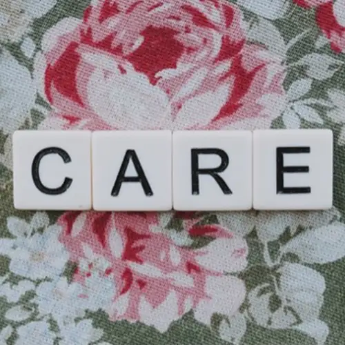 Das Wort „Care“, gelegt aus einzelnen Buchstabenblöcken auf einem grünen Tuch mit weiß-rosa Blumen.
