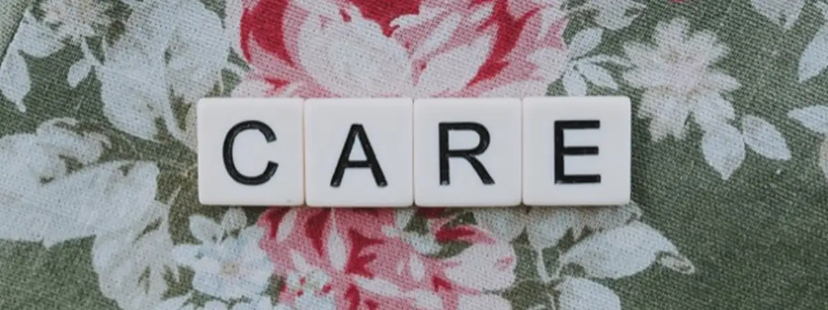 Das Wort „Care“, gelegt aus einzelnen Buchstabenblöcken auf einem grünen Tuch mit weiß-rosa Blumen.