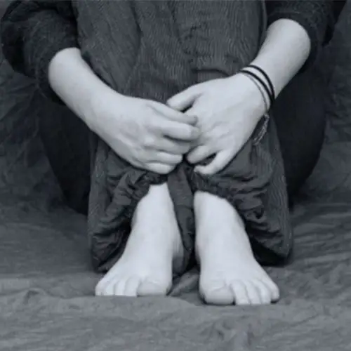 Ein schwarz-weißes Foto einer Person, die auf dem Boden sitzt und ihre Beine mit ihren Armen umschließt. Sie ist barfuß und nur der Unterkörper ist zu sehen.