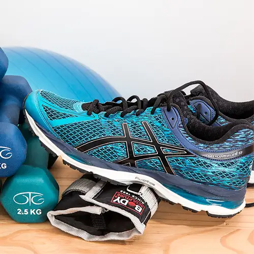 Das Bild zeigt eine Auswahl an Fitnessgeräten, darunter verschiedene Hanteln von 2 kg bis 3 kg, ein Paar türkisfarbene Sportschuhe und schwarze Gewichtsmanschetten. Im Hintergrund ist ein blauer Gymnastikball teilweise sichtbar.