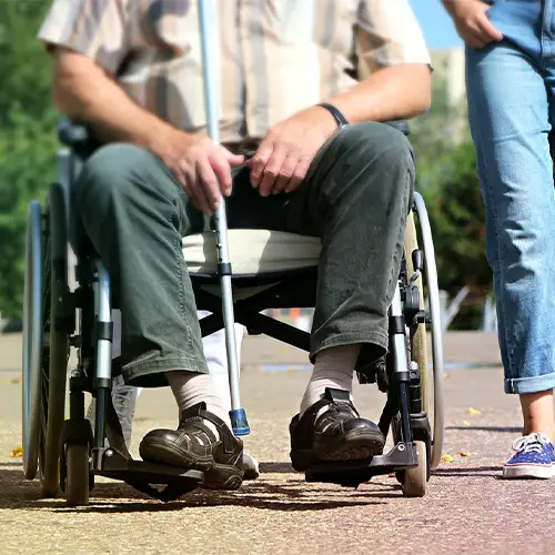 Die Beine von zwei Personen: ein Mann sitzt im Rollstuhl, eine Frau geht neben ihm.