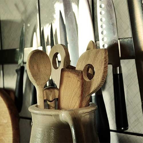 Ein Foto mit Holz-Küchenutensilien in einem Keramikbehälter vor einer Wand mit Messern an einem magnetischen Messerhalter. Daneben befinden sich zwei Schneidebretter aus Holz. 